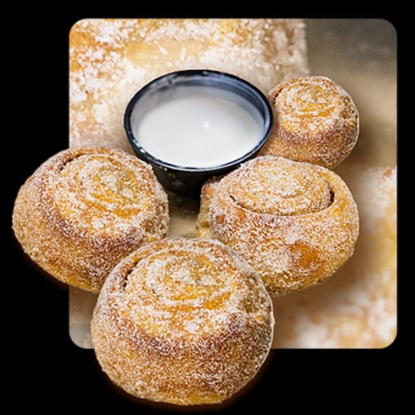 Four Golden Lizzard Pizza cinnamon rolls, dusted with sugar, surrounding a small cup of white icing.